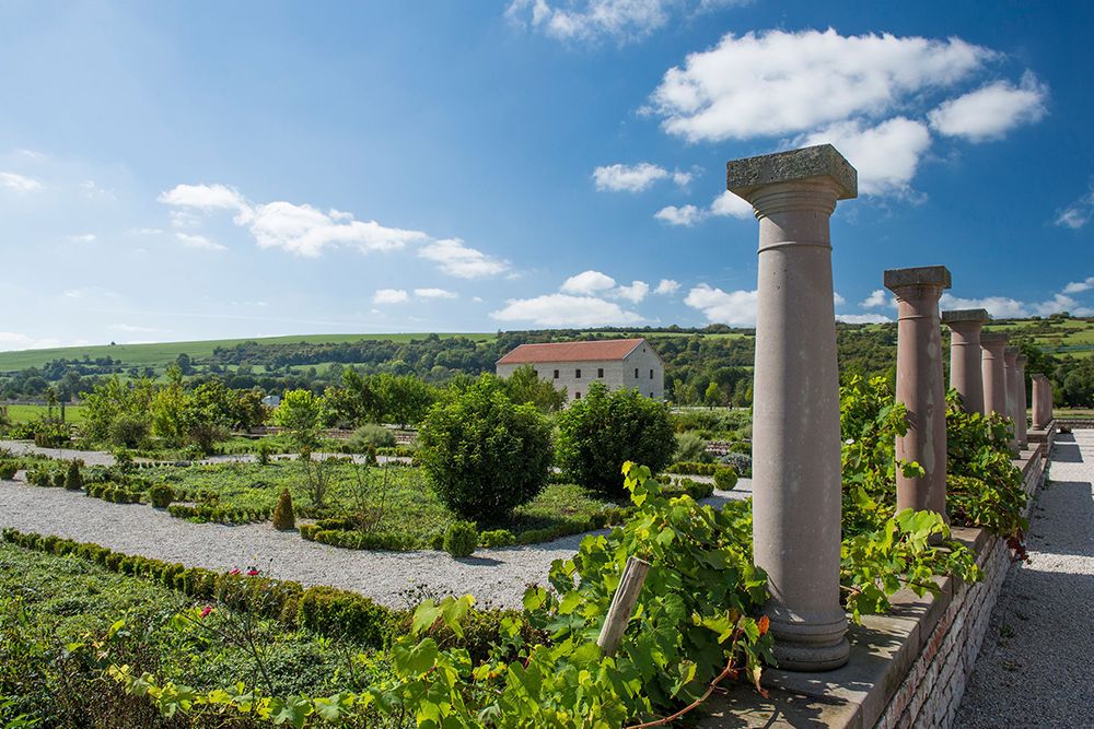 Sunny day in a garden with stone pillars and a distant building against a backdrop of rolling green hills.