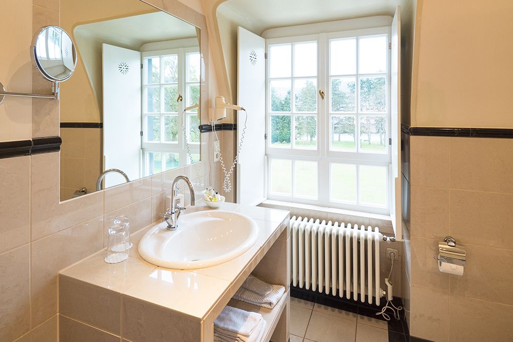 Das Badezimmer verfügt über ein Waschbecken mit Spiegel, Glas und Haartrockner. Ein Fenster sorgt für Tageslicht und darunter befindet sich ein Heizkörper.