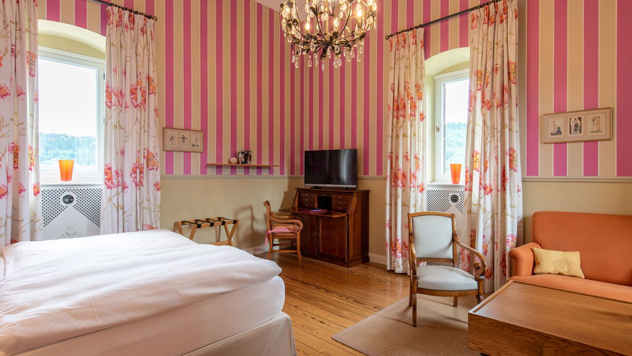 Ein Schlafzimmer mit rosa gestreiften Wänden, einem Bett, einem Fernseher auf einem Holzschreibtisch und zwei Sesseln strahlt moderne Baukultur aus. Ein orangefarbenes Sofa ergänzt die geblümten Vorhänge, während ein Kronleuchter, der an die Eleganz von Schloss Saareck erinnert, von der Decke hängt.