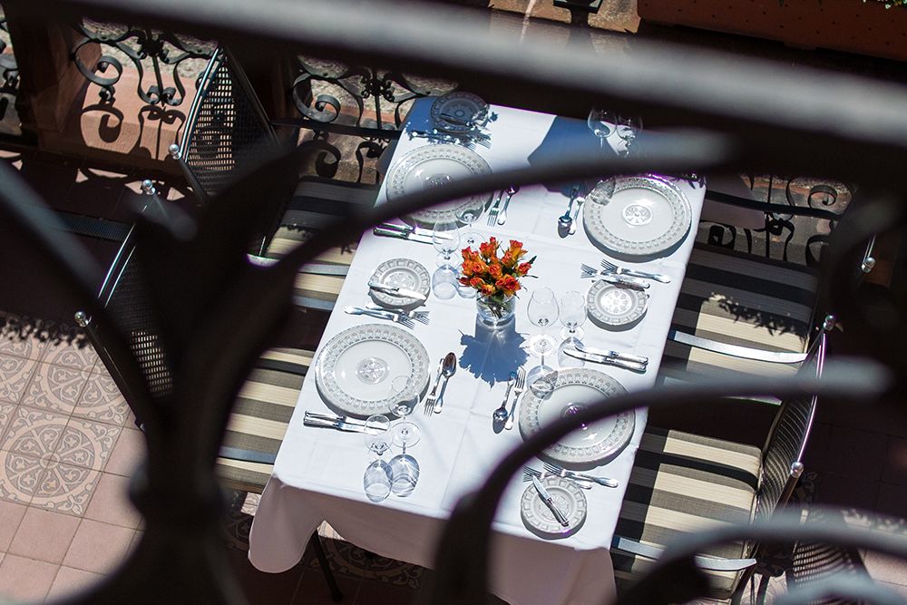 Ein für vier Personen gedeckter Tisch mit weißen Tellern, Gläsern und Besteck auf einer weißen Tischdecke. Ein kleiner Strauß orangefarbener Blumen schmückt die Mitte. Von oben betrachtet ist er durch das verzierte schwarze Geländer auf der bezaubernden Schlossterrasse mit Blick auf die friedliche Saar zu sehen.