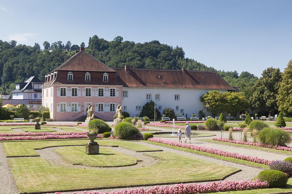 On a sunny day, a large historic building with a red roof is surrounded by manicured gardens and paths.