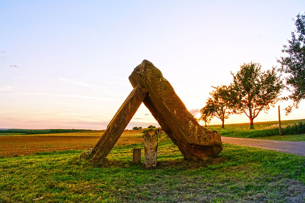 A large triangular stone structure stands on a grassy area near a road at sunset, with trees and open fields in the background. This picturesque spot is perfect for spontaneous day trips and provides an ideal base for local activities.