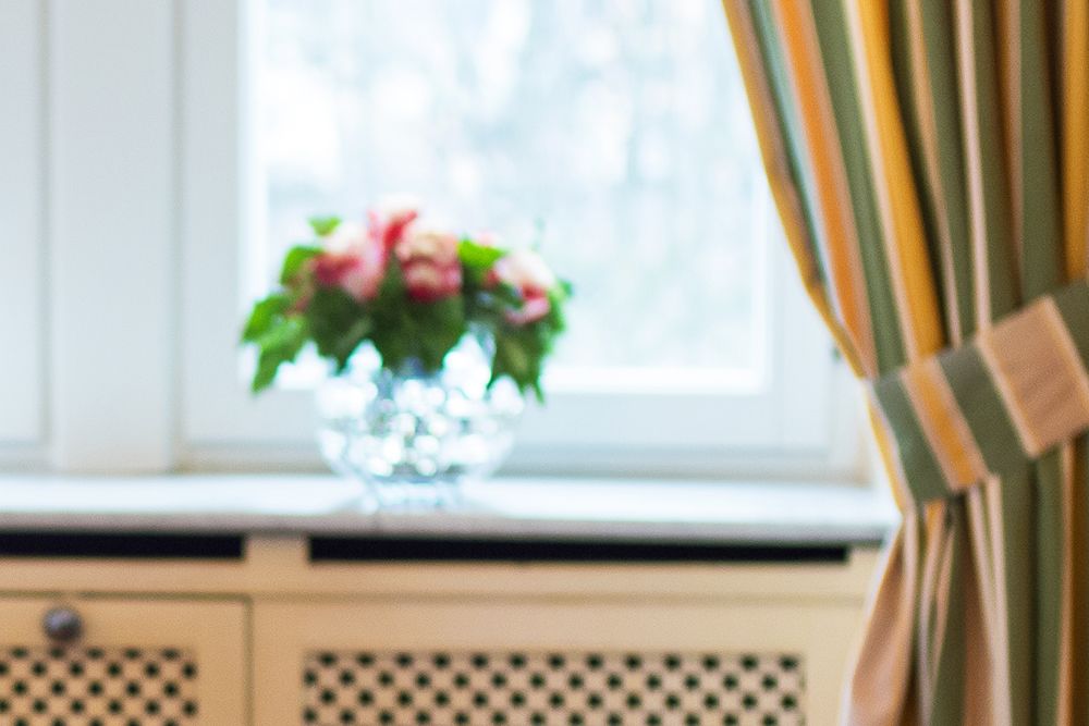 Eine Vase mit roten und weißen Blumen ziert die Fensterbank im „Gelben Zimmer“. Die gelb und grün gestreiften Vorhänge sind teilweise sichtbar.