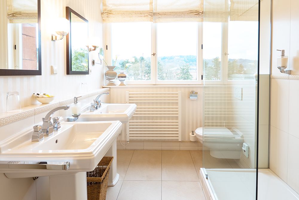 Helles Badezimmer mit Doppelwaschbecken, großem Spiegel und verglaster Dusche. Ein Fenster sorgt für Tageslicht.