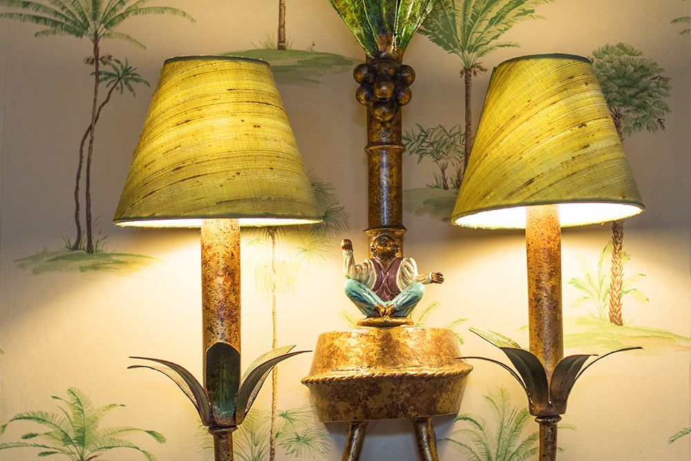 Two rustic lamps with green shades and gold bases, decorated with decorative leaves, flank a small statue of a person. The background is a conservatory wallpaper with tropical plant motifs, creating an atmosphere of subtle relaxation.