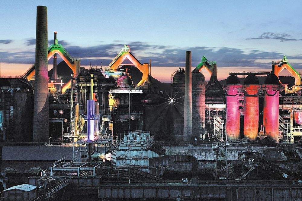 Industrial complex with brightly lit buildings, large chimneys and colourful lights at sunset under a cloudy sky.