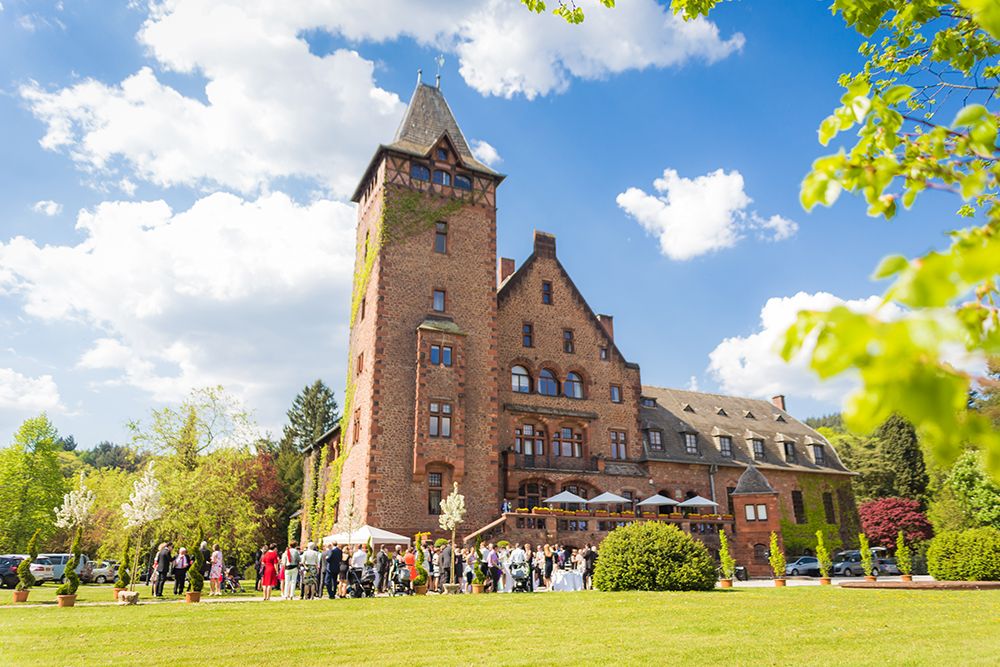 A large, historic brick building with a tower set against a blue sky, surrounded by lush greenery. A group of people gather near the entrance on a sunny day.
