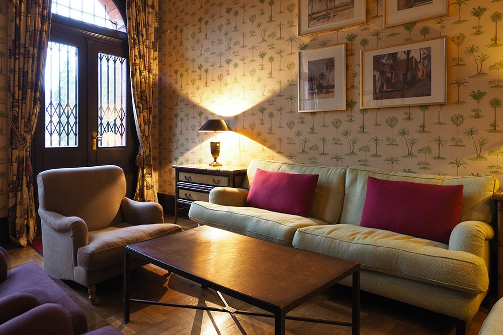 A cozy living room with a green sofa, two red pillows and an armchair offers ultimate relaxation. There is a wooden coffee table, a lamp on a side table and framed pictures on a palm-patterned wallpaper.