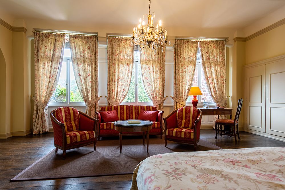 Zimmer mit geblümten Vorhängen, rot-gold gestreiften Möbeln, einem Kronleuchter und einem Schreibtisch am Fenster.