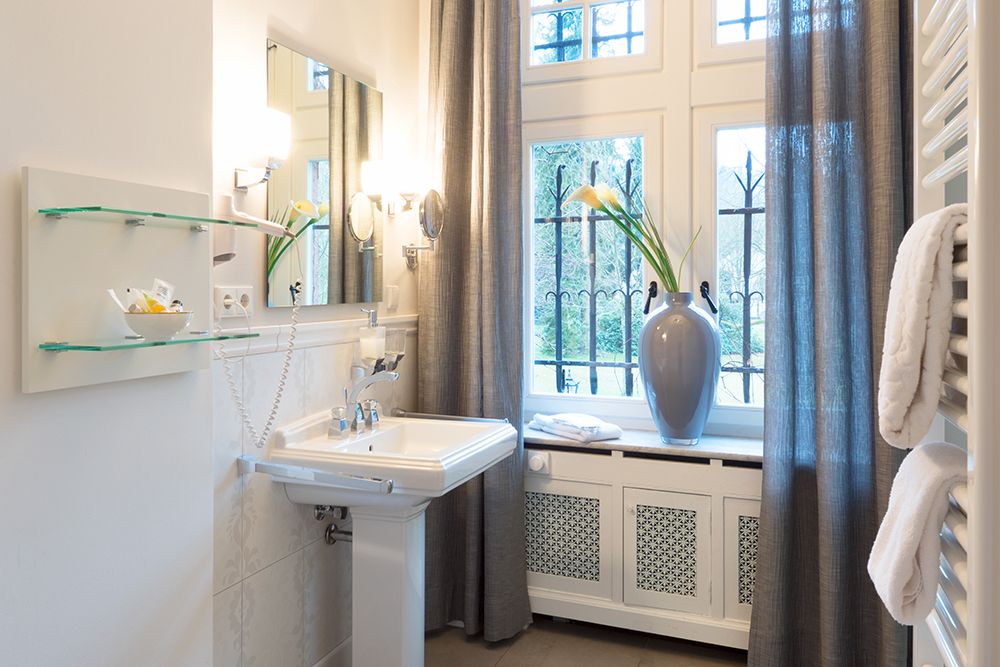 Sonnendurchflutetes Badezimmer mit weißem Waschbecken, Wandspiegel, großer Vase mit Blumen, Fenster und grauen Vorhängen.