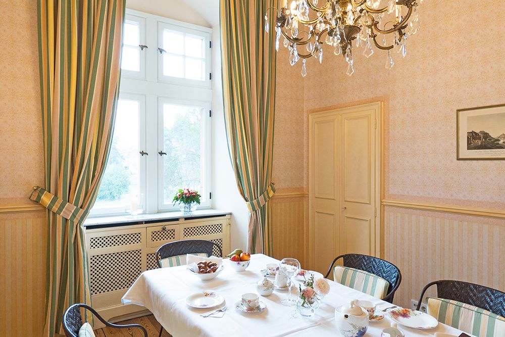 Der Speisesaal erinnert an den Charme der Alten Abtei und bietet einen mit einer weißen Tischdecke gedeckten Frühstückstisch. Stühle stehen unter einem eleganten Kronleuchter, während große Fenster mit gestreiften Vorhängen vor sanften gelben Wänden den gemütlichen Charme des Saareckparks hereinlassen.
