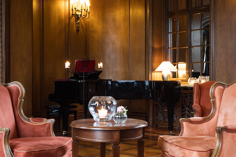 Eleganter Raum mit schwarzem Flügel, holzgetäfelten Wänden und Parkettboden. Zwei gepolsterte Stühle flankieren einen runden Tisch, der mit Kerzen und Blumen geschmückt ist. Perfekt, um in diesem gehobenen Ambiente ein Glas Single Malt Whiskey zu genießen.