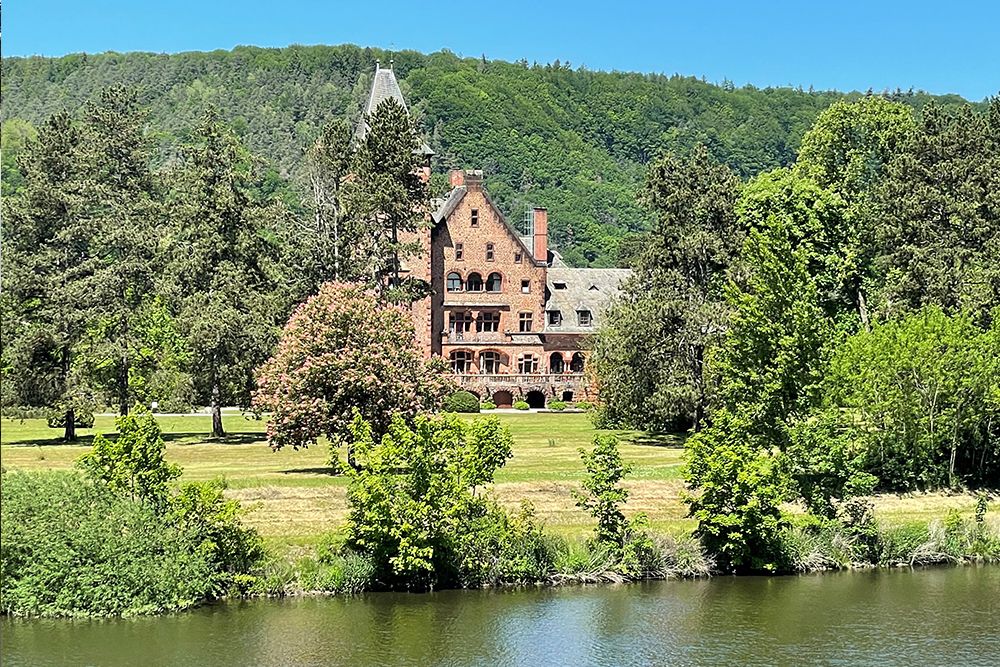 Ein großes rotes Backsteingebäude mit Spitzdach steht in einer üppig grünen Landschaft, umgeben von Bäumen, in der Nähe eines ruhigen Flusses unter einem klaren blauen Himmel.
