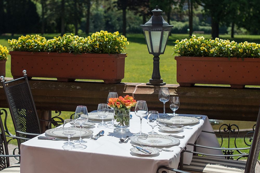 Esstisch für vier Personen im Freien mit weißer Tischdecke, Tellern, Weingläsern und einem Tischdekor aus orangefarbenen Blumen auf der Schlossterrasse. Umliegende Pflanzkübel mit gelben Blumen verleihen Charme vor dem üppigen Grün des Saarparks im Hintergrund.
