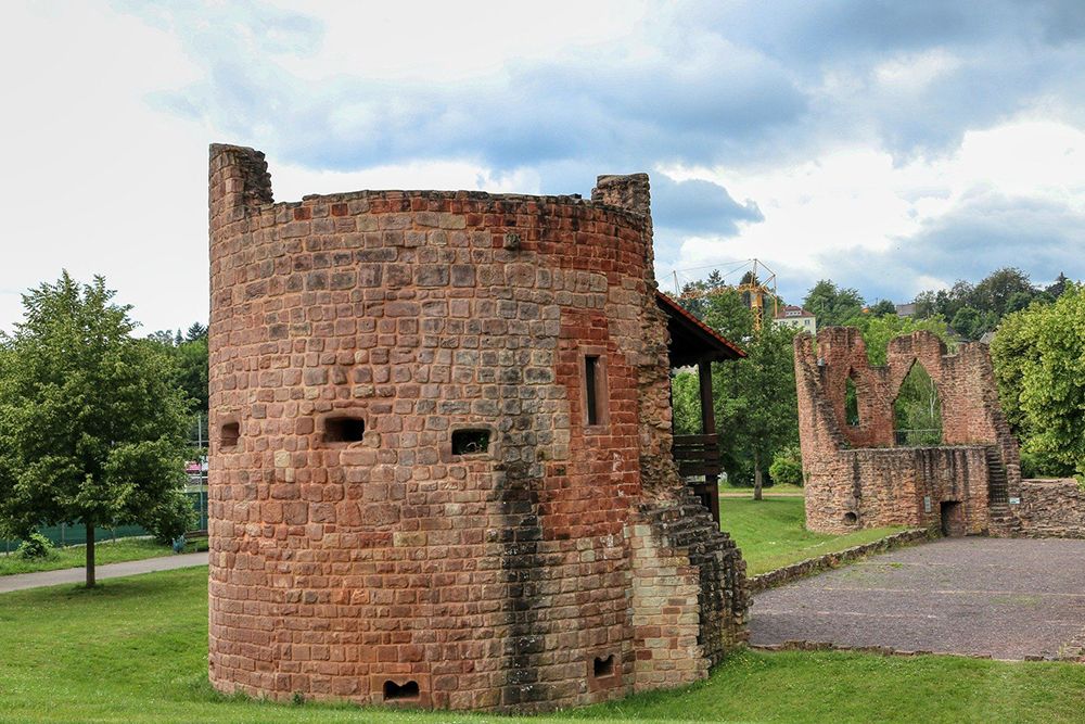 Stone castle ruins with round tower surrounded by grassy areas, trees and a cloudy sky in the background.
