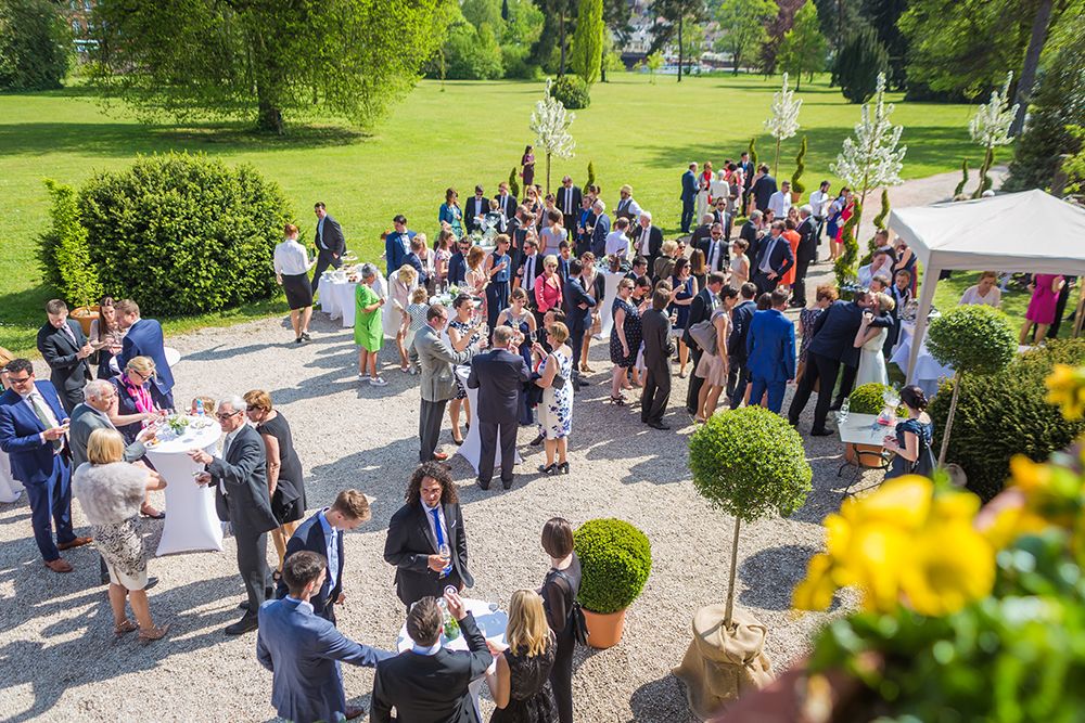 Luftaufnahme eines großen Fests auf Schloss Saareck, bei dem Gäste in formeller Kleidung gesellig zusammenkommen. Tische voller Getränke und Speisen stehen inmitten der Bäume und des Grüns des Gartens, wo alle unter freiem Himmel feiern.