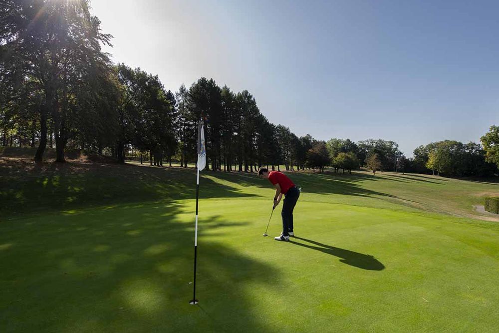 Eine Person im roten Hemd puttet auf dem sonnigen Grün eines Golfplatzes, umgeben von Bäumen und einem klaren Himmel, und verkörpert anspruchsvollen Golfsport.