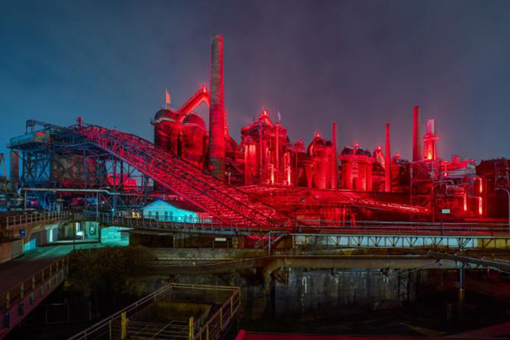 The industrial plant, a striking landmark in Saarland, glows red at night and its chimneys and metal structures stand out as a silhouette against a cloudy sky.