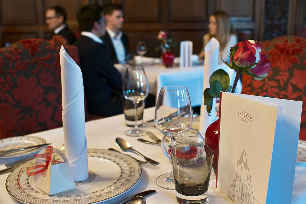 Elegant table decoration in the restaurant with folded napkins, glasses, plates and a menu. In the background you can see two couples talking. Roses in small vases decorate the tables.