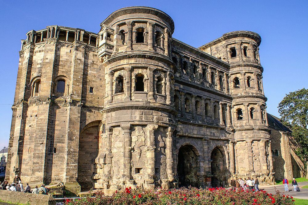 Das alte römische Tor in Trier besticht durch große gewölbte Eingänge, mehrere Ebenen und Fenster. Dieses steinerne Wunder ist von Gras und Blumen umgeben und liegt unter einem klaren blauen Himmel – ein perfekter Start für Stadttouren, bevor man sich einigen lokalen Einkaufsfreuden hingibt.