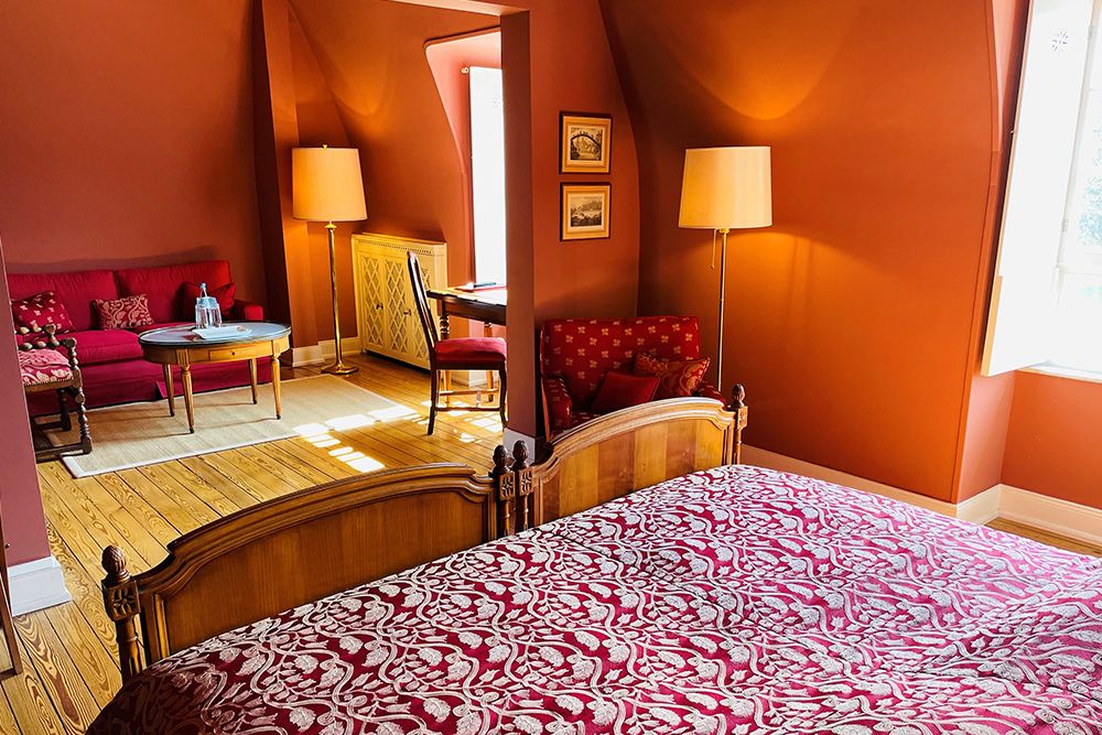 Ein warm beleuchtetes Schlafzimmer mit einem Holzbett, rot-weiß gemusterter Bettwäsche, angrenzender Sitzecke und zwei Stehlampen.