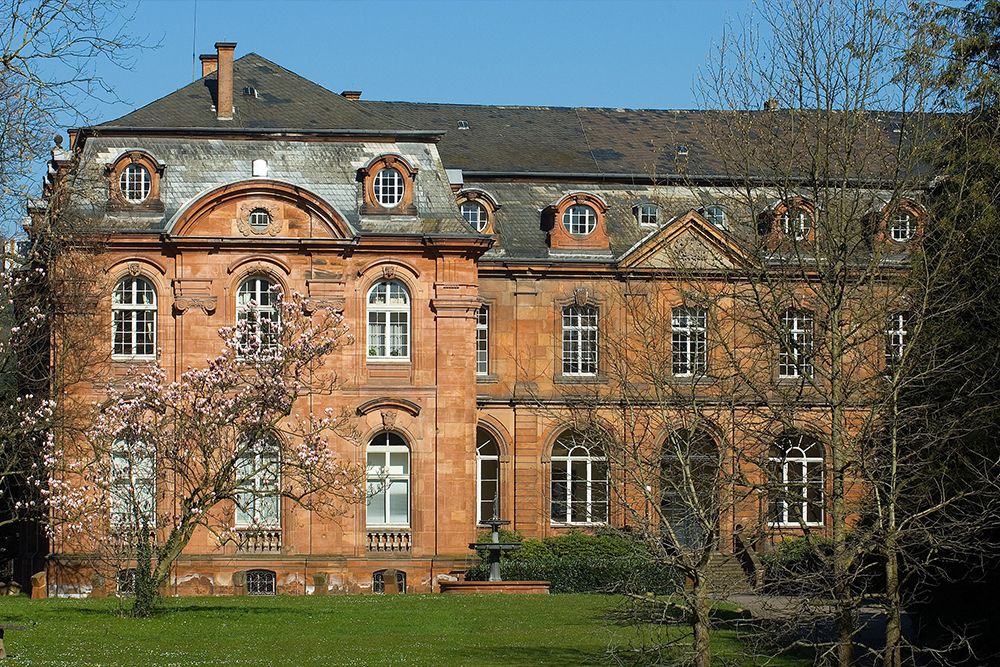 Historisches Backsteingebäude mit Bogenfenstern und kunstvollen Details, umgeben von Bäumen und einer kleinen Rasenfläche. Im Vordergrund steht ein blühender Baum unter einem klaren blauen Himmel und bietet die perfekte Kulisse für die Kammermusiktage, bei denen Musik und Kultur im Einklang mit der Natur lebendig werden.