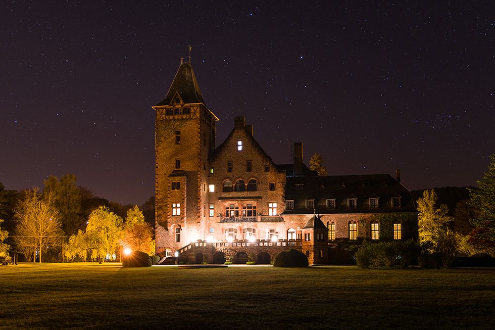 Unter einem sternenklaren Nachthimmel, umgeben von Bäumen und einer Wiese, steht ein großes, beleuchtetes Backsteinhaus mit Turm.