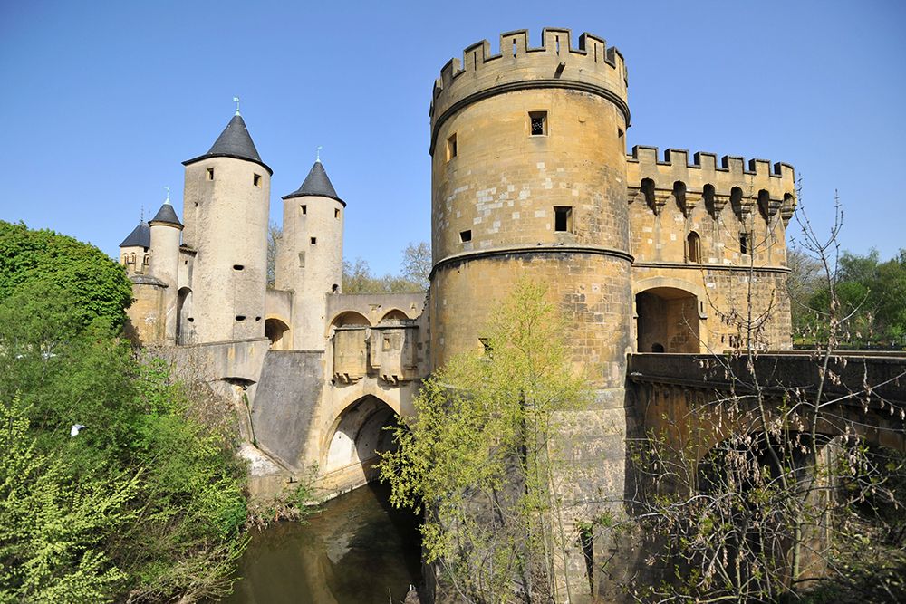 Ein atemberaubender Blick auf eine mittelalterliche Steinburg mit runden Türmen, verbunden durch eine Brücke über einen ruhigen Fluss, alles eingehüllt in üppiges Grün unter einem klaren blauen Himmel.