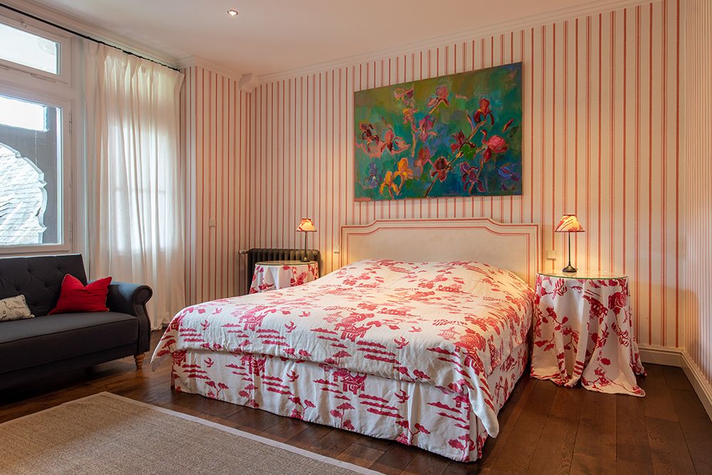 Schlafzimmer mit rot-weiß gestreifter Tapete, geblümter Bettwäsche, einem Sofa und einem farbenfrohen Gemälde über dem Bett.