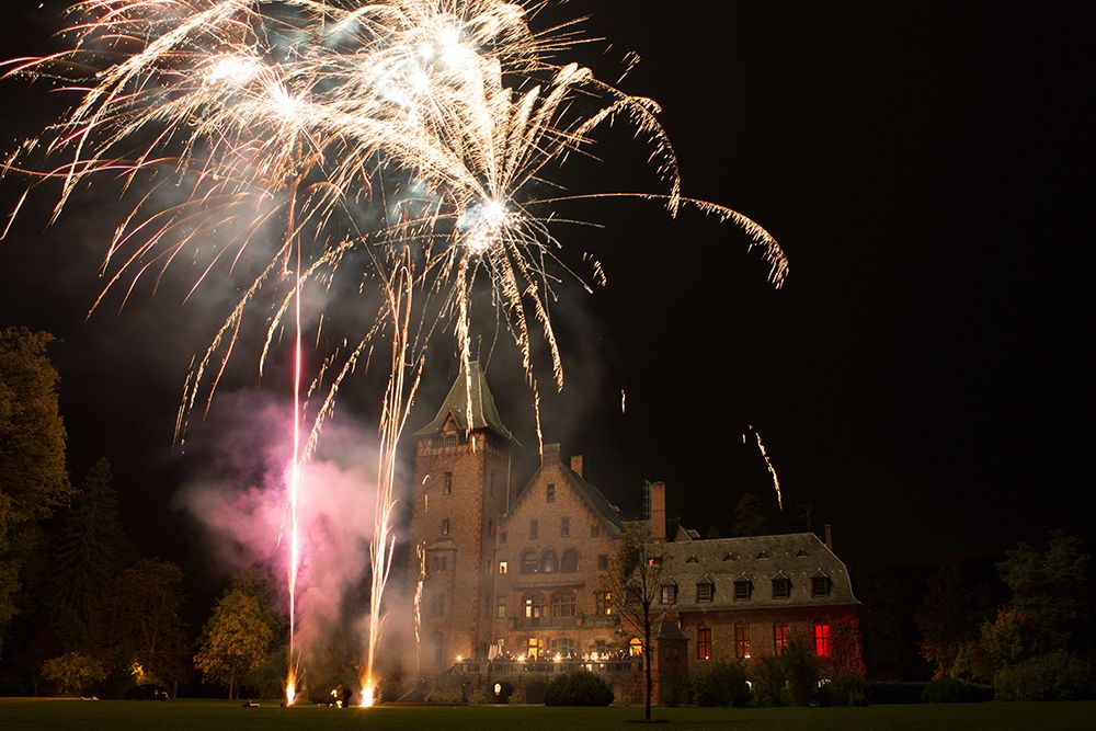 Feuerwerke erhellen den Nachthimmel über Schloss Saareck, einem großen historischen Gebäude, das von Bäumen umgeben ist, und lassen bunte Funken herabsprudeln. Es ist eine atemberaubende Erinnerung daran, dass das Leben wirklich ein Fest ist.