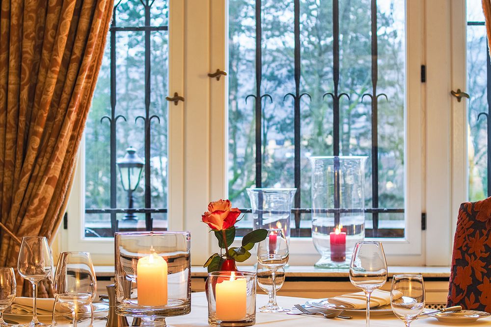 Dining table with glasses and plates, burning candles and a rose in a vase, perfect for small celebrations. Through the large window with bars, trees sway gently outside, reminiscent of a painting by Anna Boch.