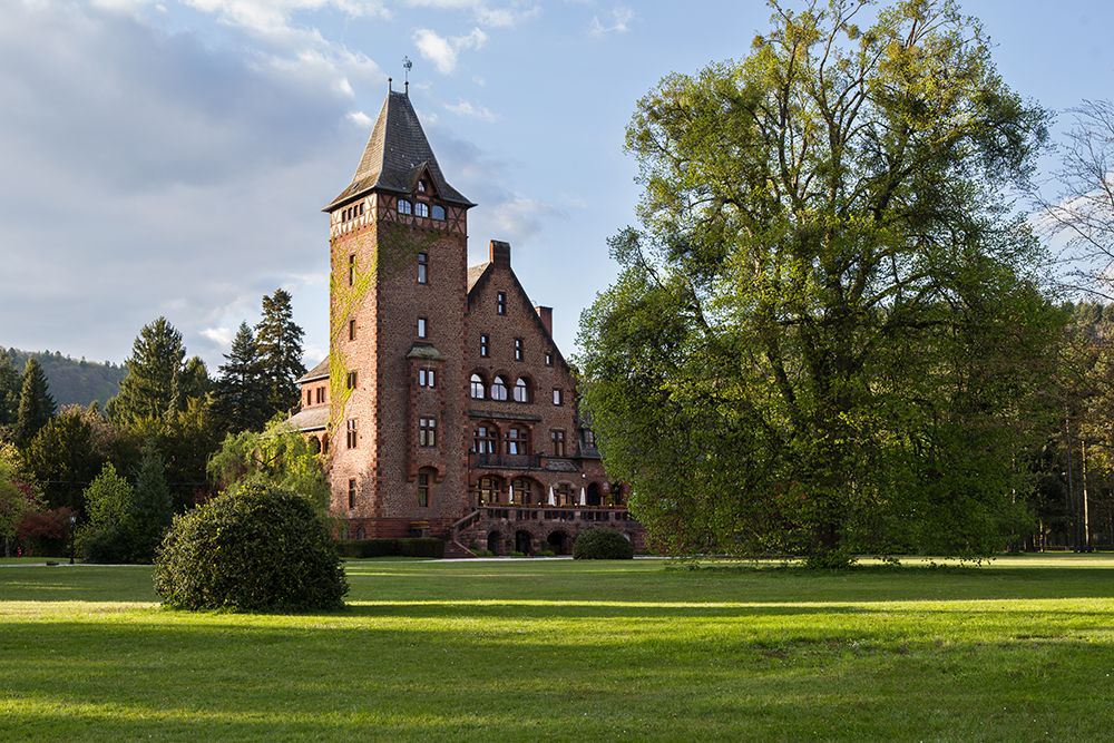 Ein großes, historisches Backsteinhaus mit einem hohen Turm steht inmitten eines grünen Rasens und Bäumen unter einem teilweise bewölkten Himmel.