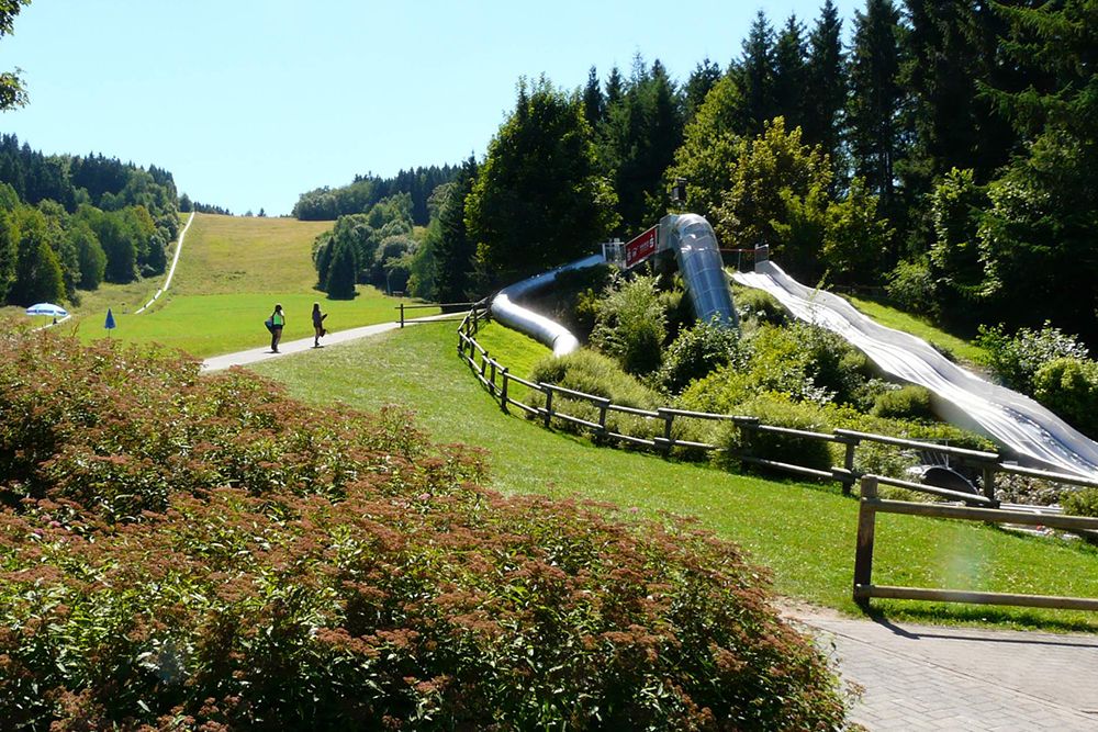 A park on a slope with a metal slide and a path. Two people walk near the slide. Trees and grass surround the area under a clear sky.