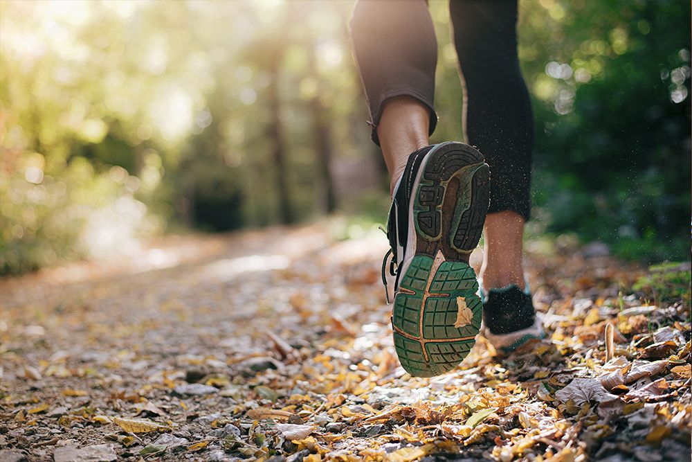 Close-up of a person jogging on a leaf-covered path, wearing black and white running shoes and black leggings, surrounded by trees and dappled sunlight. The scene captures the spirit of the sport, combining the serenity of hiking with the energy of a fast run.