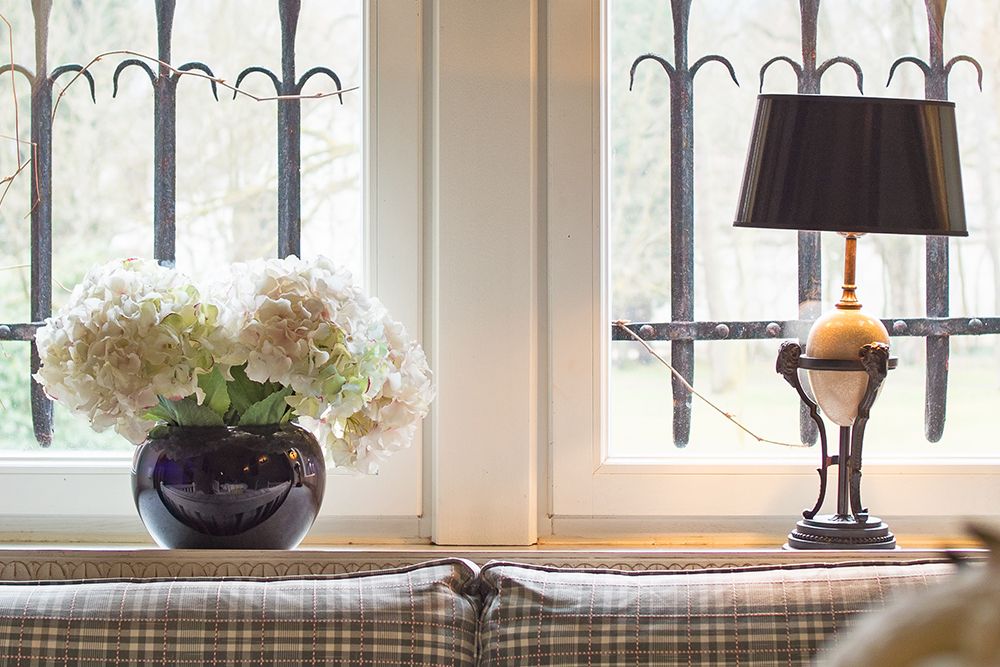 Eine Vase mit weißen Hortensien und eine dekorative Lampe zieren die Fensterbank und verstärken auf subtile Weise den Reiz der Privatsphäre mit Metallgittern, die einen zauberhaften Blick nach draußen einrahmen.