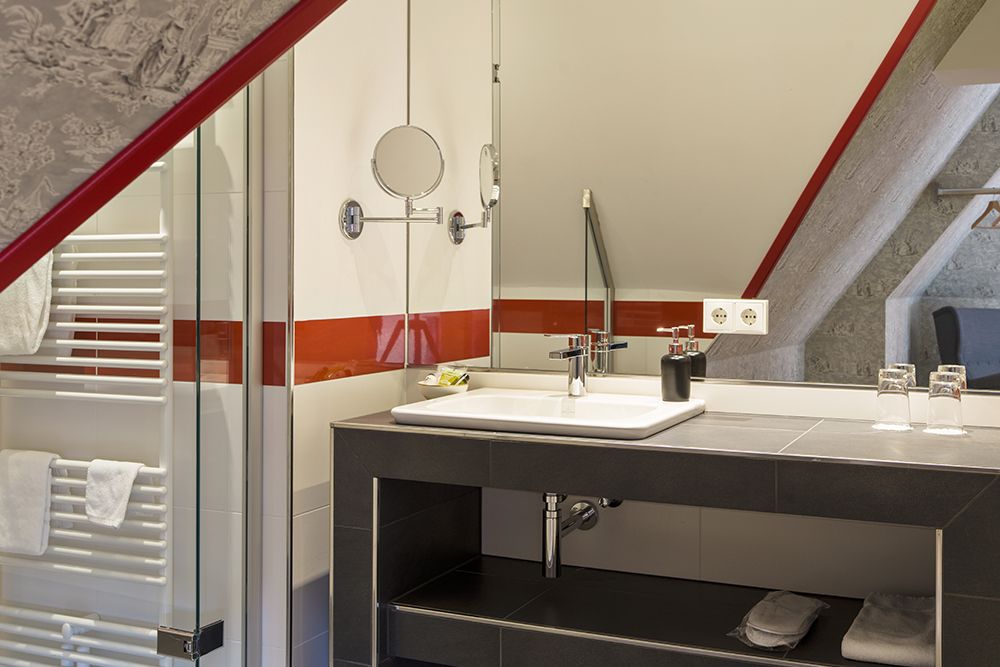 Modernes Badezimmer mit Dachschräge, Waschbecken auf dunkler Arbeitsplatte, zwei Gläsern, Toilettenartikeln, Spiegel und Heizkörper mit Handtüchern.
