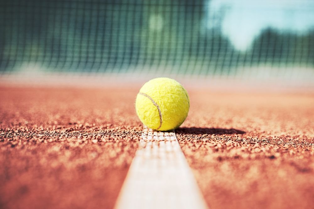 A tennis ball rests on a white line on a clay tennis court, reminiscent of the discipline and concentration required in both tennis and jogging. The net looms high in the background.