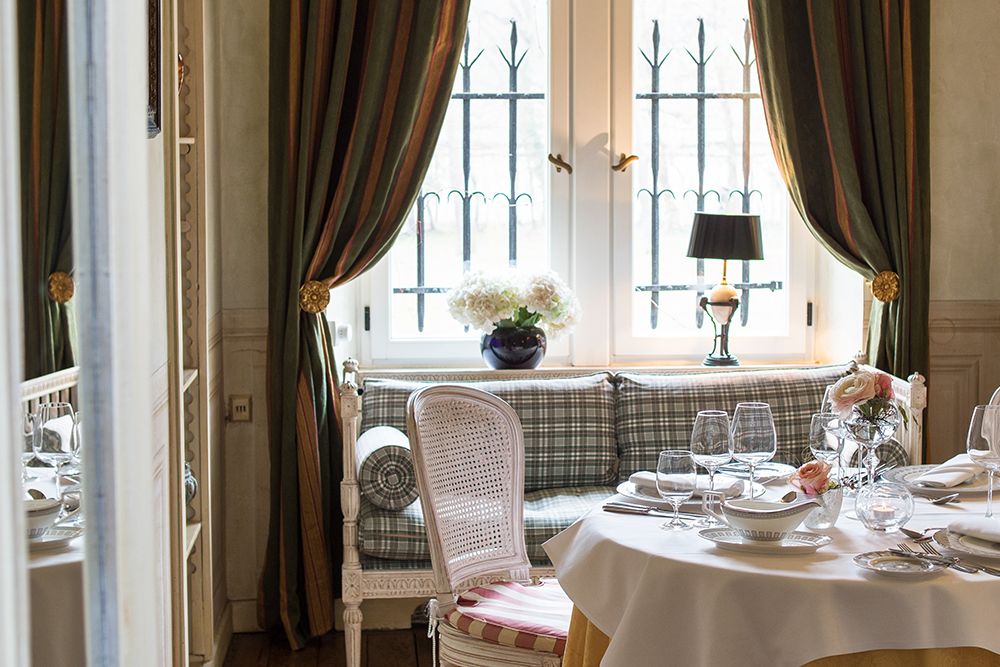 Elegantes Esszimmer mit gedecktem Tisch, perfekt für ein Geschäftstreffen oder ein gemütliches Abendessen zu zweit. Eine gepolsterte Bank mit gestreiften Vorhängen und einer Blumenvase am Fenster bietet ein wenig Privatsphäre.