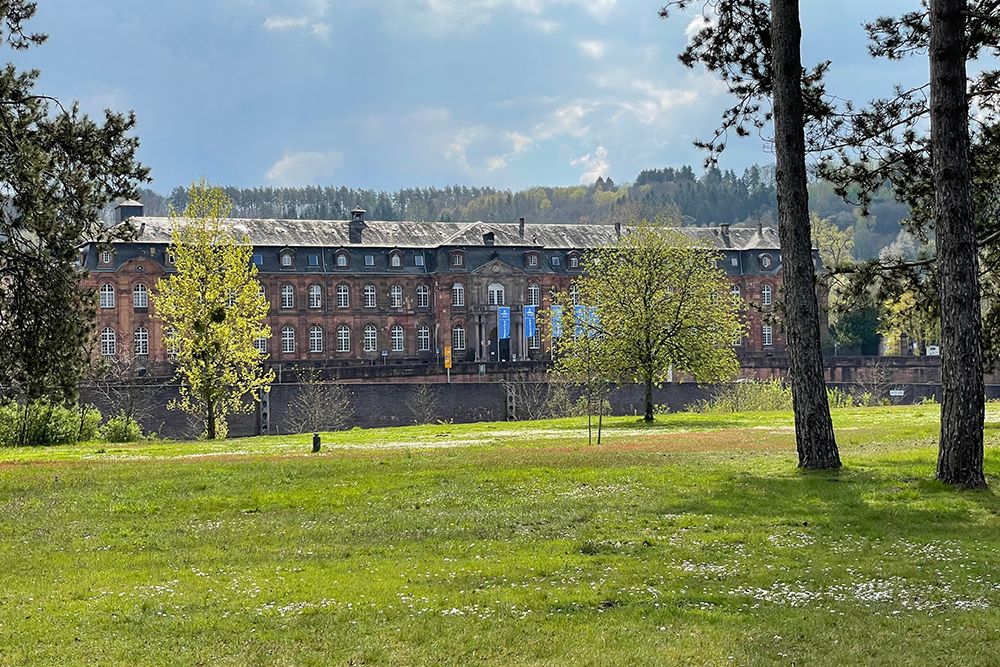 Historisches Backsteingebäude mit Bogenfenstern, umgeben von Bäumen und einer Wiese unter einem teilweise bewölkten Himmel.