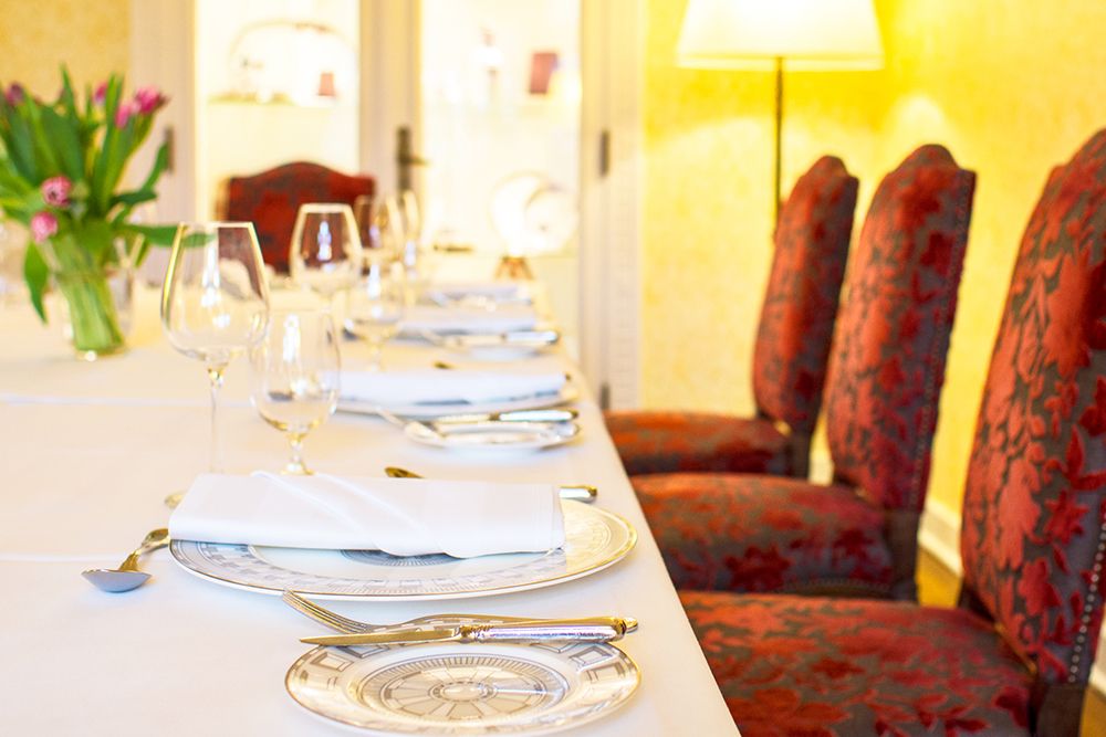 Elegant dining table set with white tablecloth, plates, cutlery and wine glasses, perfect for small celebrations. Red upholstered chairs surround the table, adding a touch of Anna Boch's artistic flair. A vase of flowers adorns the background.