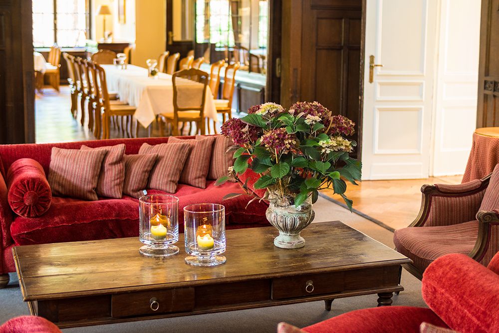 Gemütliche Hotellobby mit roten Sofas, einem Holztisch und einem Blumengesteck mit Kerzenhaltern. Im Hintergrund lädt der Speisesaal an hübsch arrangierten Tischen und Stühlen zu einem Aperitif ein.