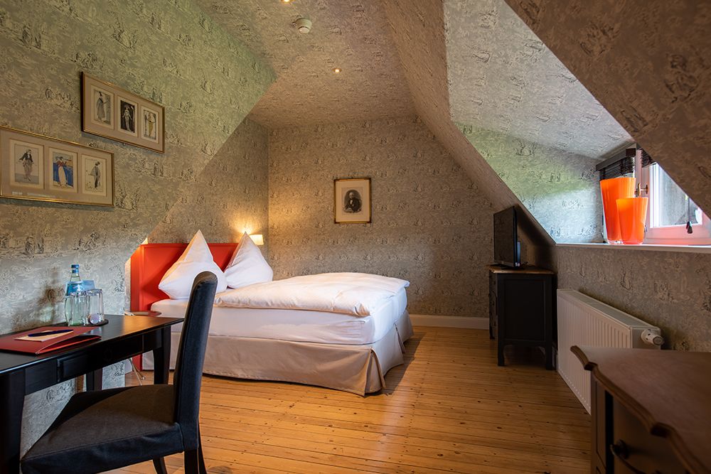 Gemütliches Schlafzimmer im Dachgeschoss mit Doppelbett, Schreibtisch, Stuhl, Wandkunst und Fernseher. Dachschräge, Holzboden und kleines Fenster.