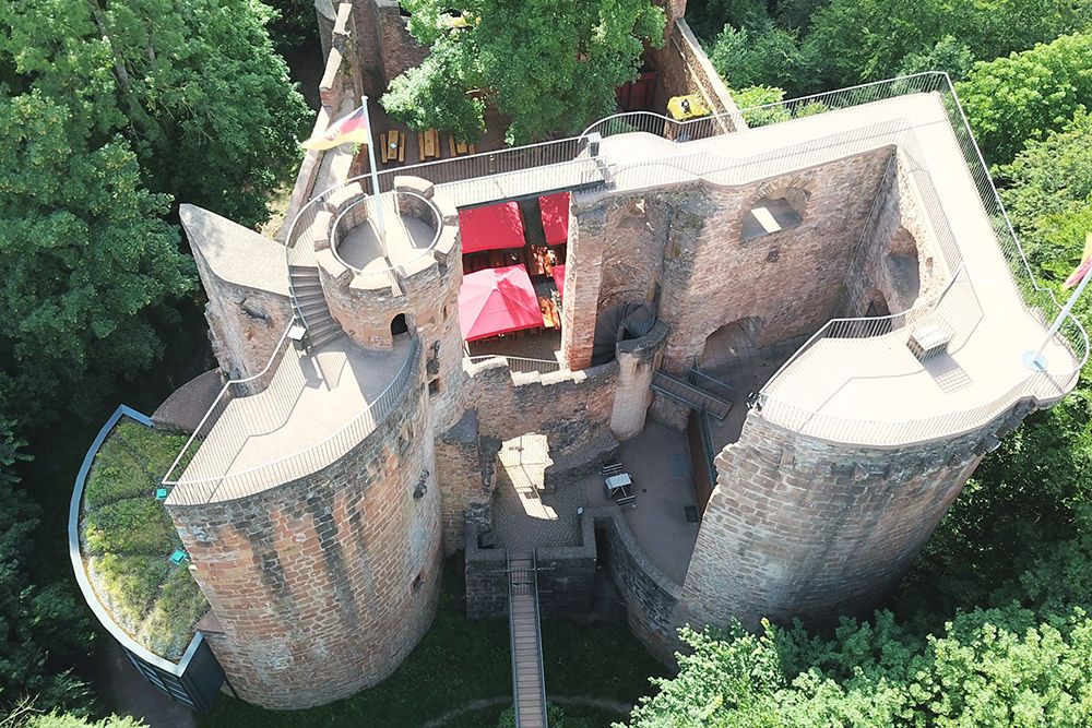 Aerial view of a historic stone castle with cylindrical towers surrounded by dense trees in Saarland. A red canopy can be seen in the castle courtyard, giving the castle charm and importance as one of the area's cherished landmarks.