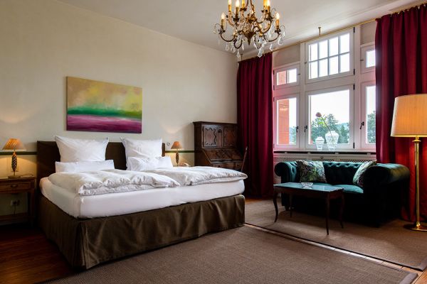 Das Hotelzimmer im Stil der Baukultur verfügt über ein Doppelbett mit weißer Bettwäsche, ein grünes Sofa und einen Holzschreibtisch mit Lampe. Ein Kronleuchter schmückt die Decke, während rote Vorhänge das große Fenster einrahmen. Ein Gemälde verleiht dem Raum Charme und erinnert an die Eleganz von Schloss Saareck.