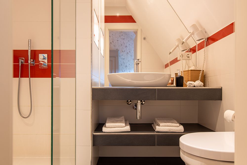 Modernes Badezimmer mit Glasdusche, roten Akzenten, weißem Aufsatzwaschbecken, Handtüchern auf Regalen und Toilette.