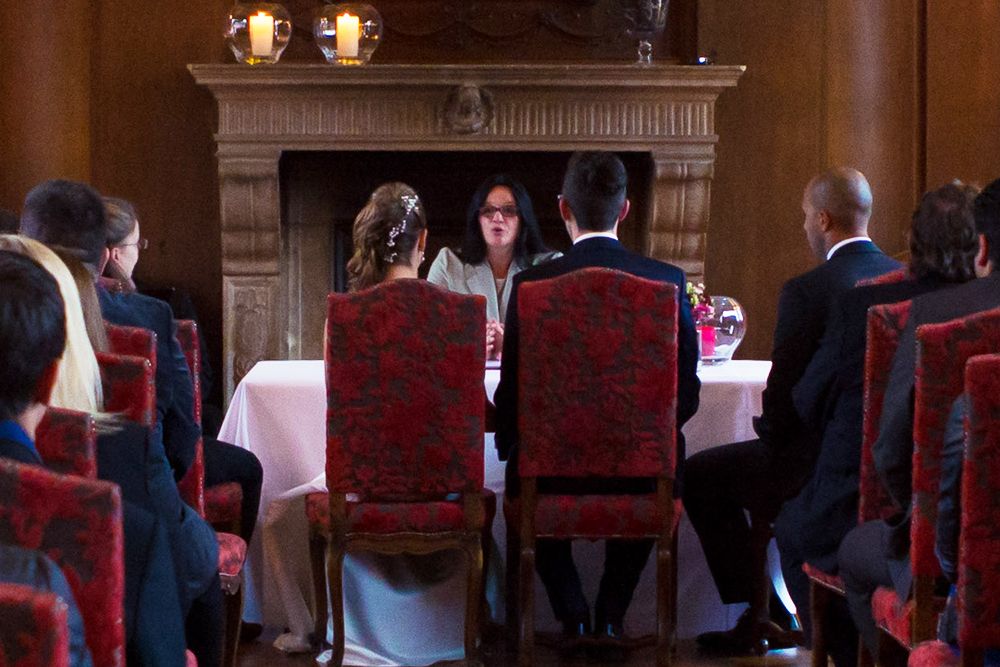 In einem bezaubernden Kaminzimmer findet eine Hochzeitszeremonie statt, bei der das Paar vor einem Geistlichen sitzt. Die Gäste sitzen auf roten Stühlen und Kerzen schmücken den Kaminsims, deren Schein durch das reiche Aroma von Single Malt Whiskey ergänzt wird, das in der Luft liegt.