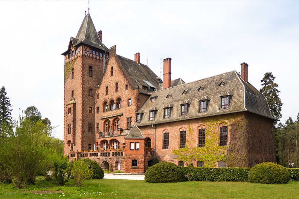 Ein großes, historisches Schloss aus rotem Backstein mit mehreren Satteldächern und einem hohen Turm, umgeben von Grün und Bäumen.