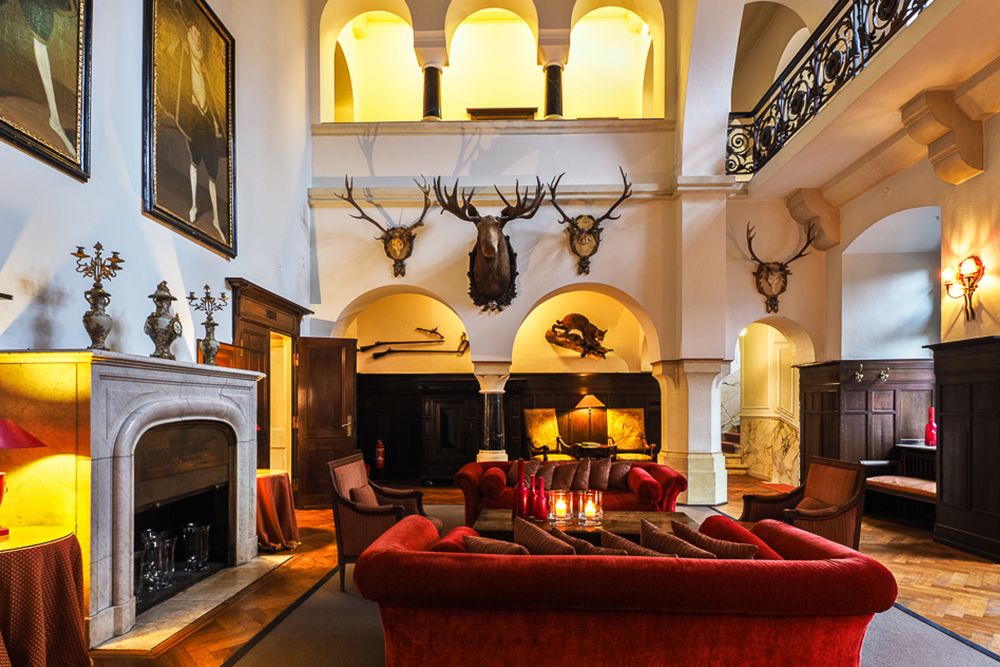 Luxuriöse Lounge mit roten Samtsofas, einem Marmorkamin und montierten Tierköpfen an hohen Wänden unter gewölbten Decken.