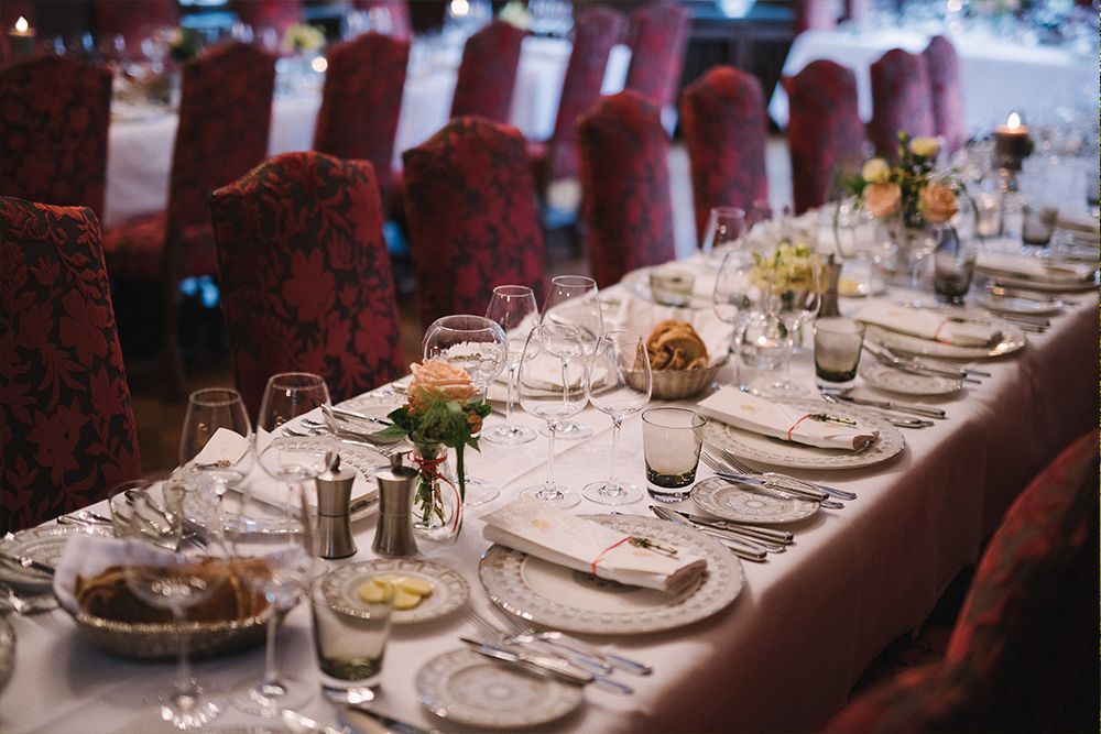 Ein eleganter Banketttisch ist mit weißen Tellern, Gläsern, Besteck und Blumendekorationen gedeckt. An beiden Seiten stehen rot gepolsterte Stühle, die den Charme einer märchenhaften Traumhochzeit hervorrufen.