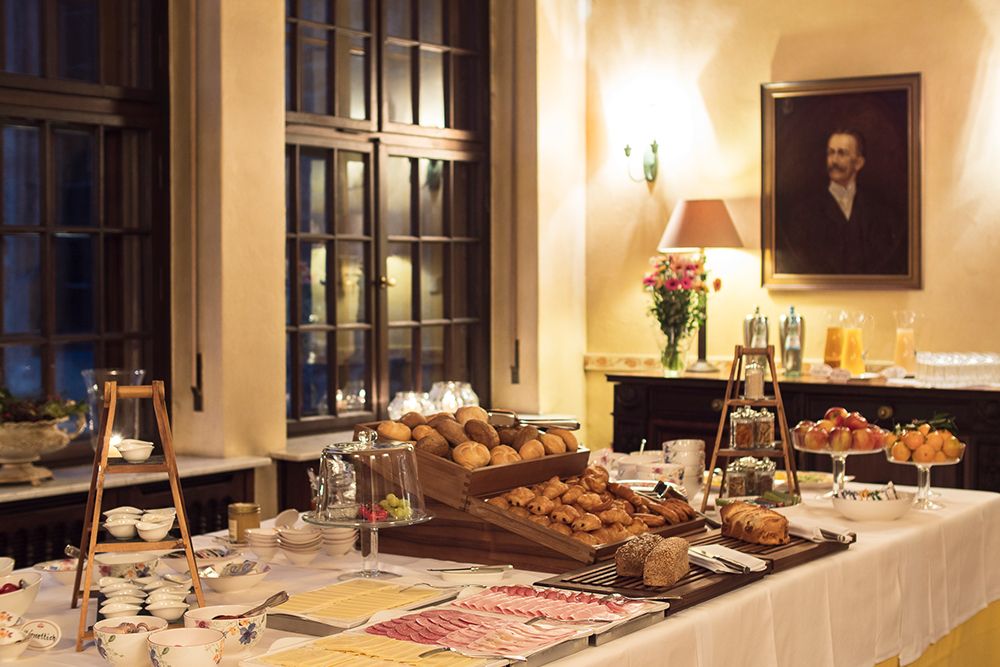 Ein Frühstücksbuffet bietet eine verlockende Auswahl an Brot, Gebäck, Fleisch, Obst und Getränken wie Kaffee und Tee. In einem warm beleuchteten Raum mit großen Fenstern und einem Porträt an der Wand lädt es die Gäste zu einem köstlichen Morgenfest ein.